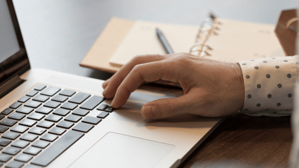 Hand typing on a laptop for content writing, with notebook and pen on a desk.
